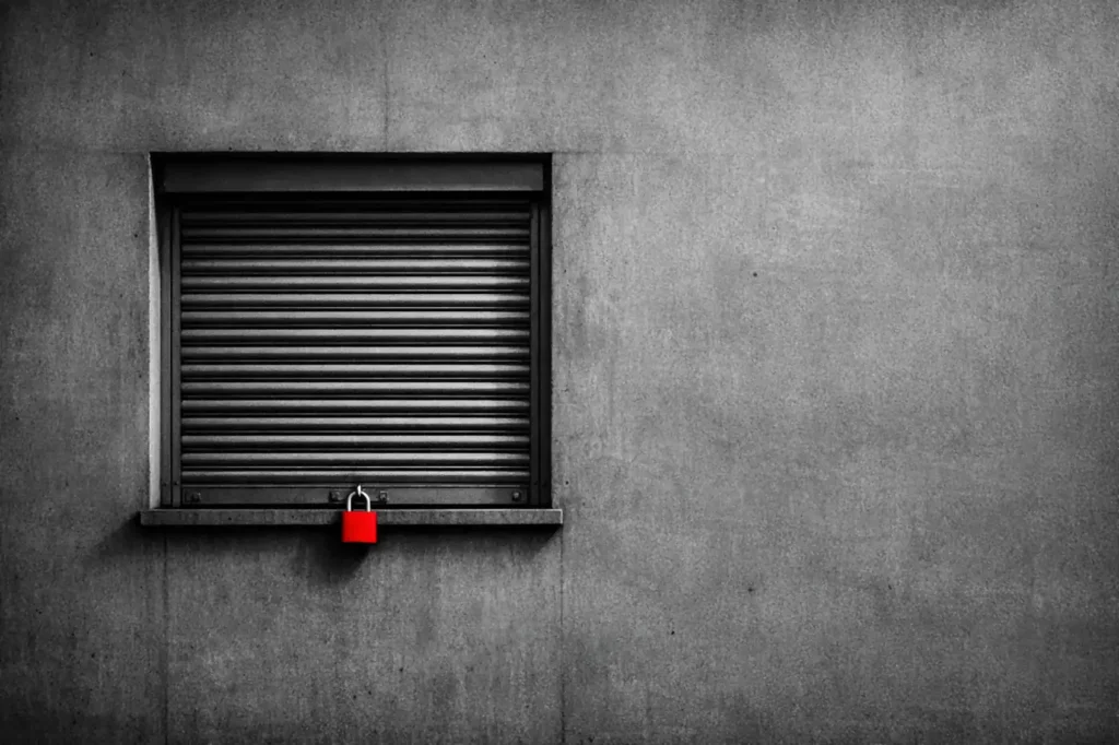 A kiosk window with its steel shutter pulled down and a red padlock hanging from the rail